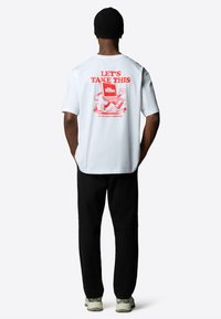 Bílý bavlněný tričko s červenou grafikou na zadní straně, na které je laptop a text. Černé kalhoty a světle barevné tenisky dokončují outfit.