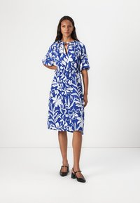 Robe florale bleue avec des motifs botaniques blancs, manches courtes bouffantes, taille froncée et encolure en forme de trou de serrure, fabriquée en tissu léger.