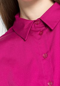 Närbild av en magenta färgad skjorta med fyra matchande knappar, som bärs av en person med ljus hy och långt brunt hår.