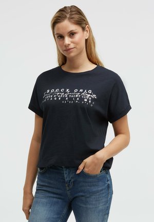 Woman with blonde hair wearing a black graphic t-shirt with rolled-up sleeves and blue jeans, hand in pocket, standing against a plain background.