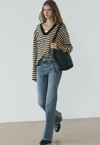 Žena chodí uvnitř, má na sobě béžový a černý pruhovaný svetr s výstřihem do V, modré džíny, černé boty a nese velkou černou kabelku přes rameno.