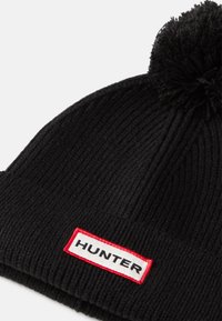 Căciulă tricotată neagră, cu o textură ribbed și un pompon în vârf. Dispune de o etichetă albă coaptă cu un bordură roșie pe care scrie "HUNTER".