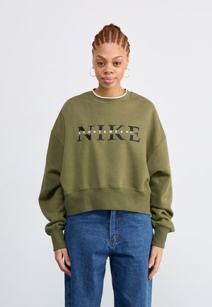 Olive green sweatshirt with a loose fit, featuring a prominent black "NIKE" logo and white "SPORTSWEAR" text, paired with blue denim jeans.
