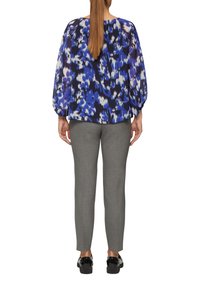 Blouse multicolore avec un motif abstrait bleu, violet et blanc ; coupe ample, manches ballons. Associée à un pantalon tailleur gris et des chaussures noires.