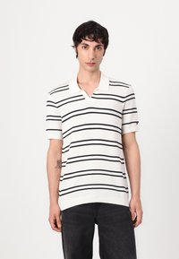 Hvit stripete polo-skjorte med svarte linjer, kort erm, og V-hals krage. Vevd tekstur; kombinert med mørke jeans.