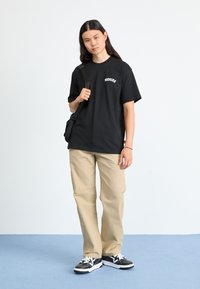 T-shirt noir à manches courtes avec texte blanc, pantalon beige et baskets noires avec accents blancs, se tenant sur un sol bleu contre un fond uni.