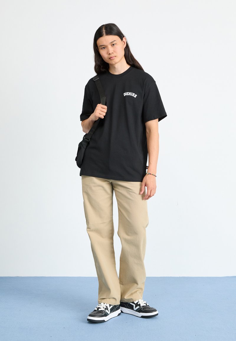 T-shirt noir à manches courtes avec texte blanc, pantalon beige et baskets noires avec accents blancs, se tenant sur un sol bleu contre un fond uni.