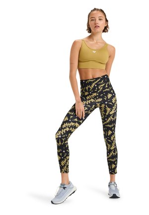 Outfit sportivo con reggiseno senape e leggings neri con motivi floreali dorati. Le scarpe da ginnastica sono di un grigio chiaro con suole scure.