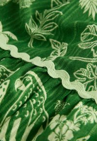 Grüner Stoff mit einem botanischen Muster aus hellgrünen Blättern und Vögeln. Wellenförmiger, hellgrüner Rand am Saum. Weiche Textur.