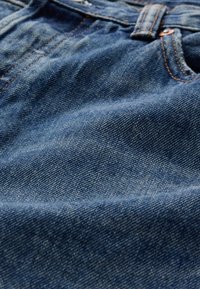 Tissu en denim bleu foncé avec une texture montrant un léger effet délavé, comprenant des coutures orange et une passants de ceinture visible.