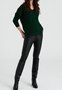 Dunkelgrüner Strickpullover mit V-Ausschnitt, getragen zu engen schwarzen Lederhosen und kniehohen schwarzen Stiefeln mit Absatz. Der Stoff wirkt strukturiert.