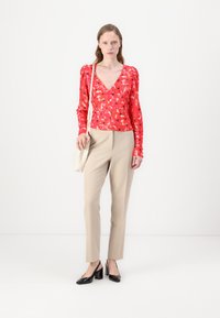 Haut à manches longues rouge à fleurs avec décolleté en V et épaules bouffantes, associé à un pantalon beige ajusté et des chaussures à talons noires.