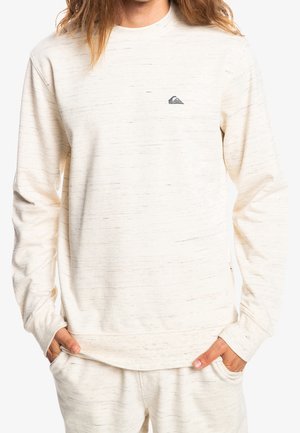 Mand iført cremefarvet langærmet sweatshirt og matchende bukser med subtilt sort spraglet mønster og lille logo på brystet.