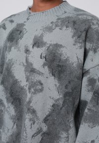 Lys grå sweater med en blød tekstur, prydet med et marmoreret mørkegrå mønster, rund halsudskæring og let slidte kanter.