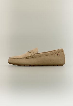 Chaussure beige de style mocassin en matériau suédé, avec une semelle plate, une couture décorative et un petit patch logo multicolore sur le côté.