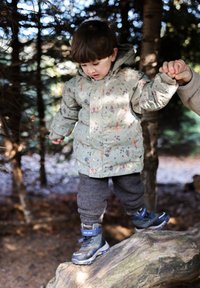 Dítě v světle zelené bundě s květinovým vzorem, šedých kalhotách a modrých nepromokavých botách stojící na pařezu v zalesněné oblasti.