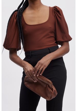 Person trägt ein braunes Oberteil mit Puffärmeln und schwarze Hose, hält eine passende braune Leder-Clutch auf Taillenhöhe.