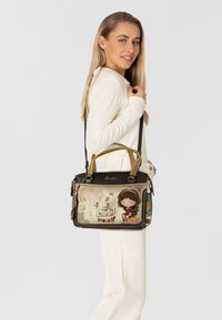 Schwarze und beige Handtasche mit einer dekorativen Illustration eines Mädchens und Text. Mit zwei Griffen, einem Schultergurt und einzigartigen Verzierungen.