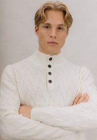 Hvit kabelstrikket genser med høy hals, med knappestolpe og mørke knapper. Teksturen er myk og ribbet.