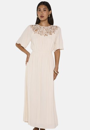 Femme portant une longue robe crème à manches courtes avec un décolleté floral découpé complexe, debout, une main sur la hanche.