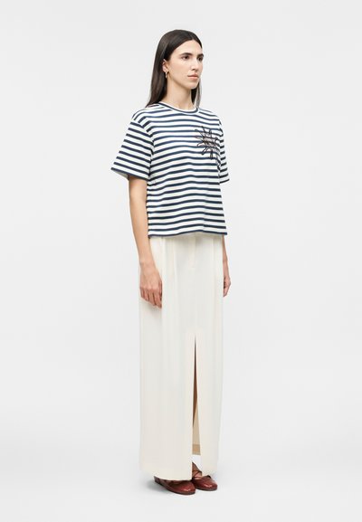 Γυναίκα που φοράει μπλε-σκουρό ναυτικό και λευκό ριγέ κοντομάνικο τοπ με κέντημα ήλιου, λευκή μακριά φούστα με σκίσιμο μπροστά και καφέ φλατ παπούτσια, στεκόμενη πλευρικά.