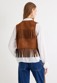 Gilet in suede marrone con frange sull'orlo, indossato sopra una blusa bianca. L'outfit presenta una vestibilità comoda e jeans in denim blu.