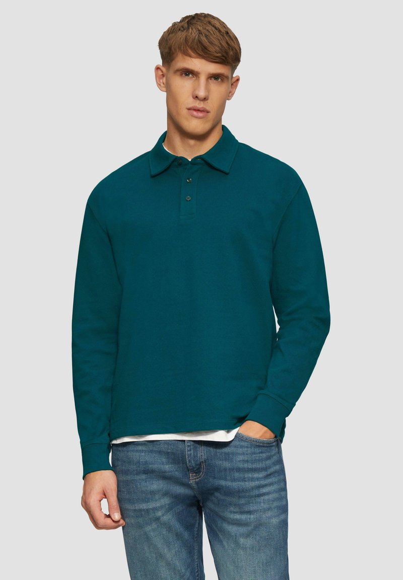 Polo shirt teal à manches longues avec col et patte de boutonnage à trois boutons, fabriqué en tissu texturé. Porté avec un jean bleu clair, les mains dans les poches.