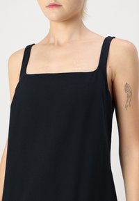 Robe noire sans manches avec un décolleté carré. Tissu lisse, design ajusté, sans motifs visibles et une légèreté subtile. Tatouage sur le bras visible.