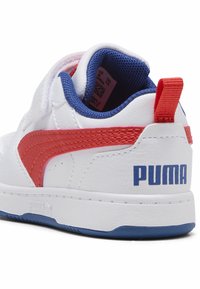 Zapatillas blancas sintéticas con acentos rojos y azules, con un acabado texturizado, cuello acolchado y la marca "PUMA" en negrita en el talón.