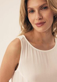 Biała bezrękawnikowa bluzka o delikatnym połysku, miękkiej teksturze i zaokrąglonym dekolcie, z delikatnymi fałdami na ramionach.