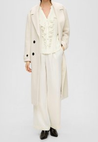 Completo in tre pezzi color avorio: cappotto lungo texturizzato, blusa con volant e pantaloni a gamba larga. Tutti i capi sono realizzati in tessuti morbidi; bottoni neri e stivali a punta.