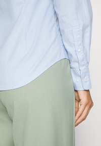 Helle blaue Button-up-Bluse mit langen Ärmeln, die einen abgerundeten Saum hat, kombiniert mit weichen grünen, weit geschnittenen Hosen, die eine glatte Textur aufweisen.