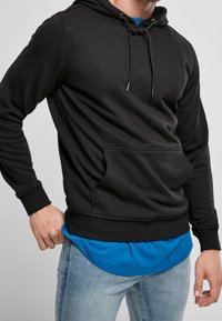 Czarna bluza z kapturem wykonana z mieszanki bawełny, z kieszenią kangurką, sznurkami z metalowymi końcówkami i ściągaczami na rękawach, nałożona na niebieską koszulkę.