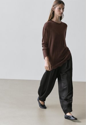 Kobieta chodząca w pomieszczeniu, ubrana w luźną brązową bluzkę z długim rękawem, szerokie czarne spodnie i czarne płaskie buty na jednolitym jasnym tle.