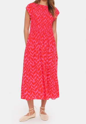 Woman wearing a red and pink patterned midi dress with short sleeves and beige lace-up woven flats, standing against a white background.