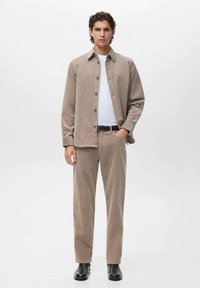 Chaqueta de camisa de pana color marrón claro con cierre de botones, combinada con pantalones marrón claro a juego. Camisa blanca debajo, cinturón negro y botas negras hasta el tobillo.