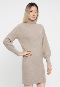 Abito a maglia a coste beige con collo alto e maniche lunghe leggermente arricciate. Tessuto morbido, lunghezza fino a metà coscia e vestibilità comoda.