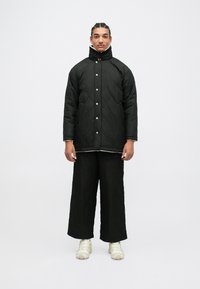 Giacca nera trapuntata con colletto alto e bottoni a pressione, abbinata a pantaloni neri a gamba larga e sneakers chiare. Texture liscia.