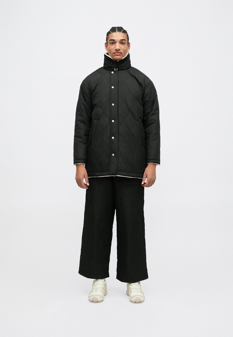 Giacca nera trapuntata con colletto alto e bottoni a pressione, abbinata a pantaloni neri a gamba larga e sneakers chiare. Texture liscia.