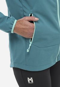 Tannengrüne Softshelljacke mit kontrastierenden mintfarbenen Reißverschlüssen, Reißverschlusstaschen an den Seiten und elastischen Bündchen. Schwarze Leggings mit weißem Millet-Logodetail.