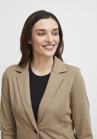 Beige blazer med slagkrage, enkelskapsknäppning och slät textur. Bärs över en svart topp, vilket skapar en koordinerad outfit.