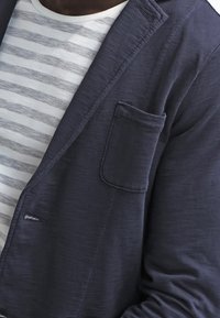 Persona con una camisa a rayas grises y blancas debajo de un blazer azul oscuro con un bolsillo en el pecho, vista de cerca.