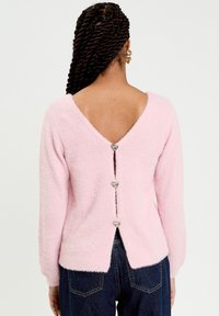 Vaaleanpunainen, pörröinen neulepaita syvällä V-mallisella selkäosalla, jossa on kolme sydämenmuotoista nappia ja sivuhalkiot. Pitkät hihat ja pehmeä pinta.