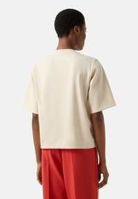 Krémově zbarvený top s krátkými rukávy, rovného střihu, hladkou texturou a jednoduchým výstřihem, ve spojení s rezavě zbarvenými širokými kalhotami.