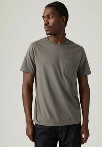En mand med fletninger modellerer en grå t-shirt med en brystlomme og mørke jeans, mod en lys neutral baggrund.