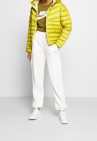 Měkká světle zelená bunda s kapucí, hnědé tričko s bílým logem, bílé tepláky a bílé tenisky s jemnými akcenty.