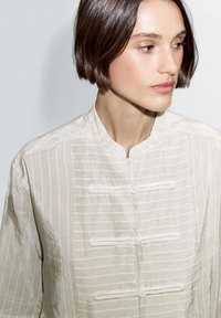 Jasnobeżowa, długi rękaw bluzka z kołnierzem mandaryńskim, z pionowymi paskami, zapięciami na guziki oraz fakturowaną tkaniną.