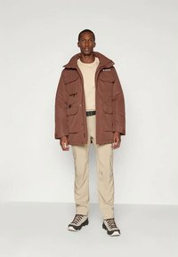 Veste brune isolante avec capuche, plusieurs poches avant et fermeture éclair. Associée à une chemise légère et un pantalon cargo beige.