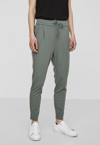 Grå joggerbyxor med dragsko i midjan, avsmalnande ben och veckdetaljer. Bärs med vita sneakers och en svart topp.