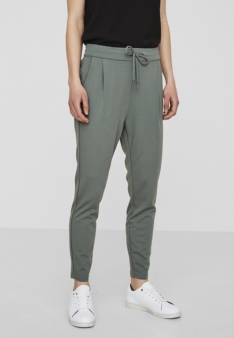 Grå joggerbyxor med dragsko i midjan, avsmalnande ben och veckdetaljer. Bärs med vita sneakers och en svart topp.